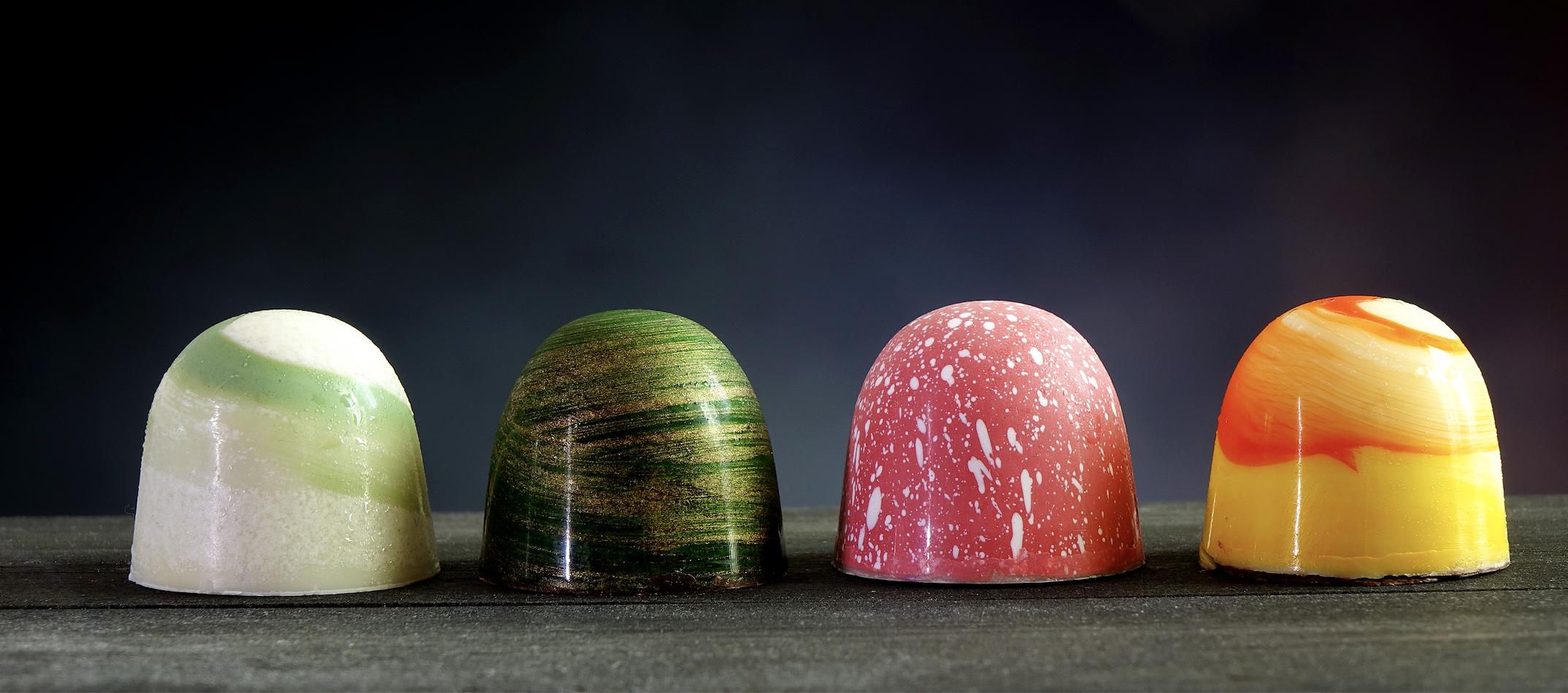 A professional display of four gourmet chocolate bonbons with vibrant, glossy coatings in red, yellow, and orange, achieved with BioconColors high-stability natural colorants.